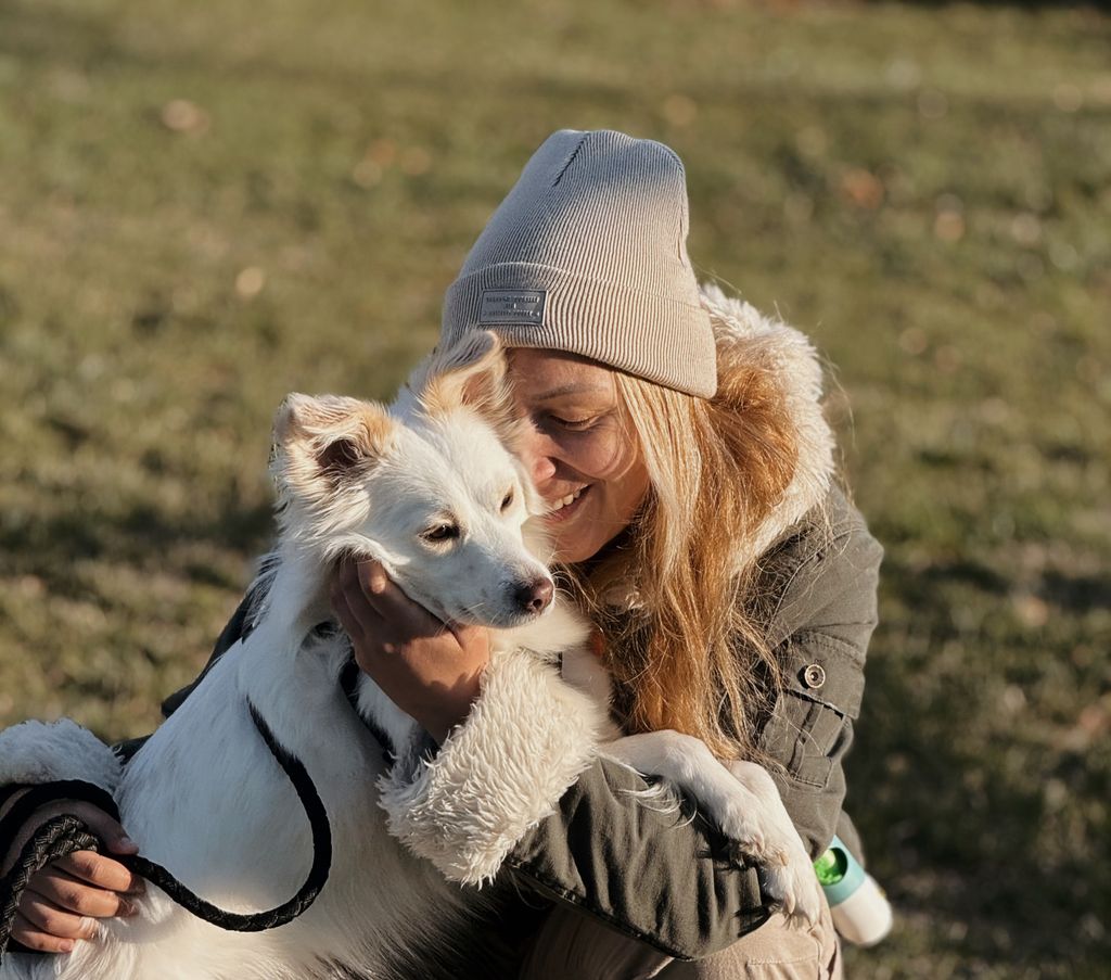 A woman in a beanie snuggles with her white mixed breed dog in a sunlit outdoor setting.